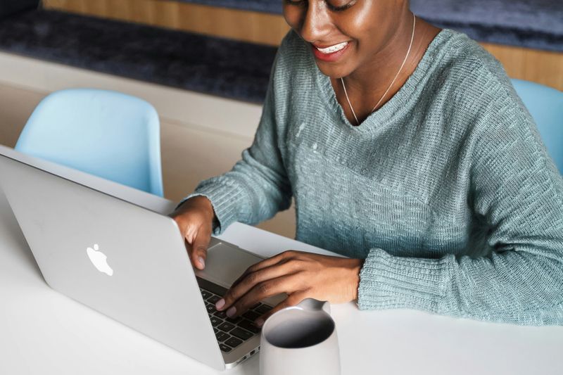 A woman is sitting with a laptop smiling. Beside her is a cup.