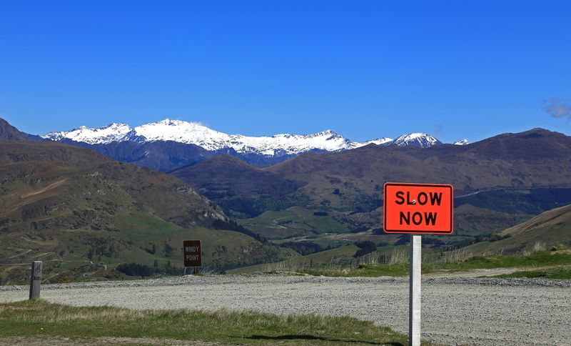 An orange sign in front of a mountain road that reads 'Slow Now.'