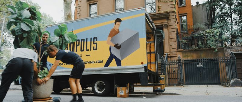 Three movers carry a potted plant towards the back of a moving truck.