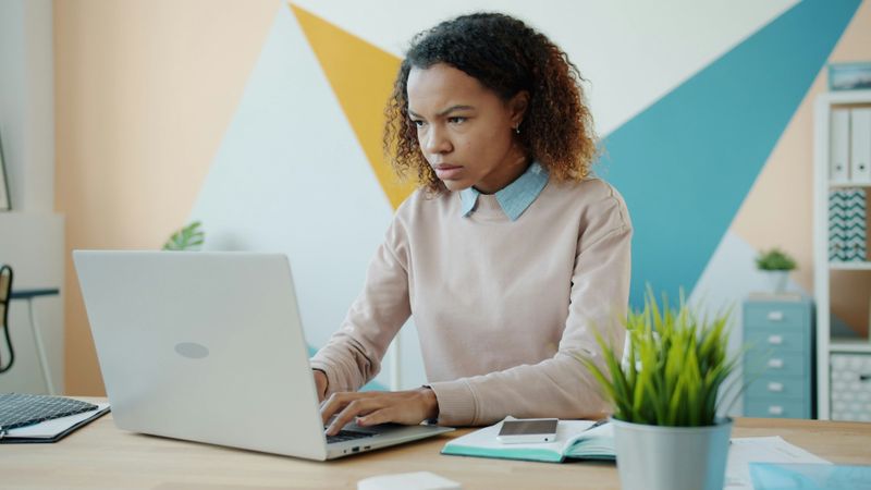 A woman sitting in a workplace and looking at the laptop screen with a confused face. 