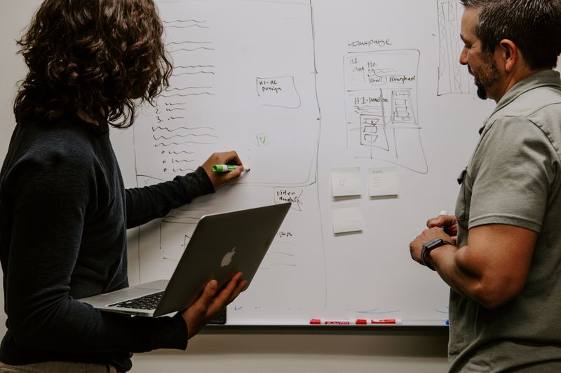 Two coworkers in front of a whiteboard. One coworker holds a laptop in one hand and writes on the board with a marker.