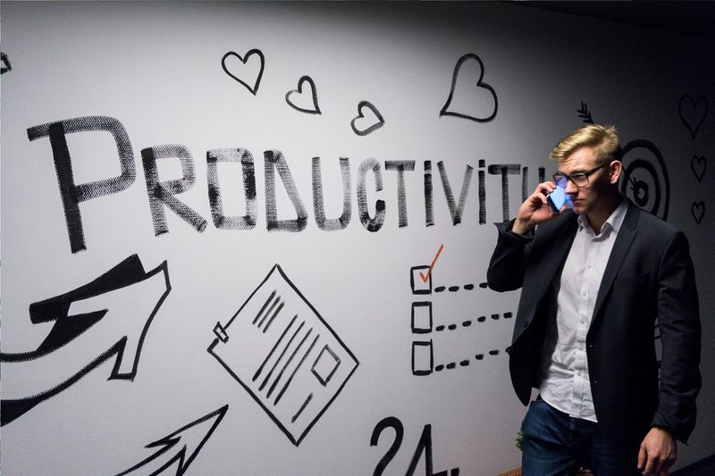 A man in glasses and blazer on a phone. He stands in front of a mural that depicts a productive workday.