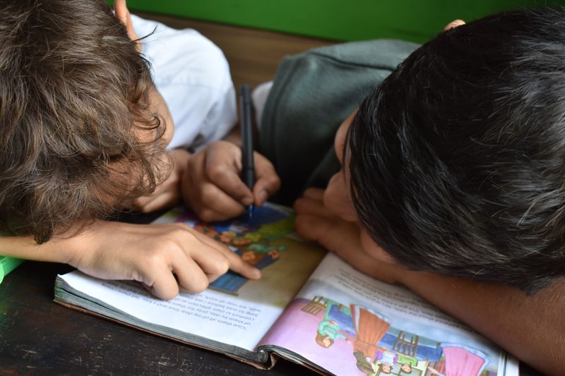 Two boys reading a book together, while resting their heads on either side of the book. One boy is holding a pen.