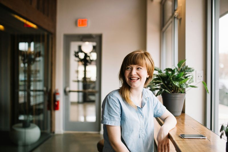 A woman sitting by a window, smiling. 