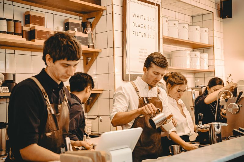 5 workers in aprons behind a counter prepare coffee orders in front of a wall of coffee items and menu