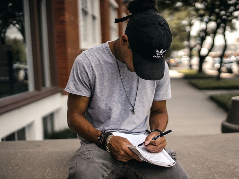 Image of a young man  wearing a cap, sitting outside and writing in a notebook.
