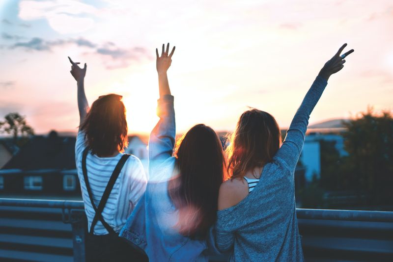 Photo of three friends, from behind, holding up hands while facing a sunset.