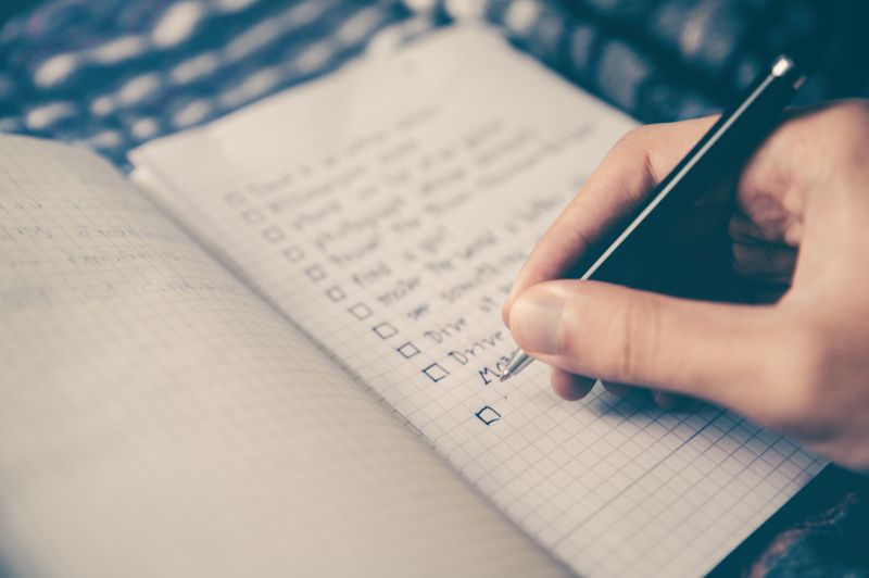 A hand holding a pen writing a list of TO DO items in a notebook.