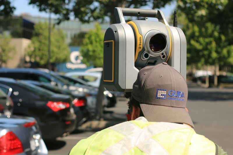A surveyor bending down to look through surveying equipment in parking lot.