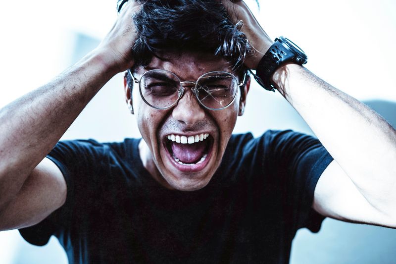 A guy is grabbing his hair, shutting his eyes, and screaming with his mouth wide open. One side of his glasses is cracked. 