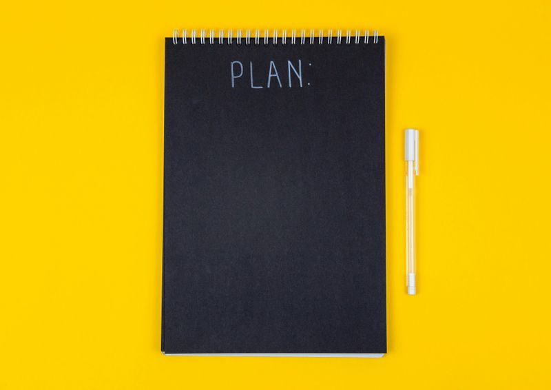 A black spiral notebook with “PLAN:” written at the top lies on a yellow background, with a pen placed beside it.