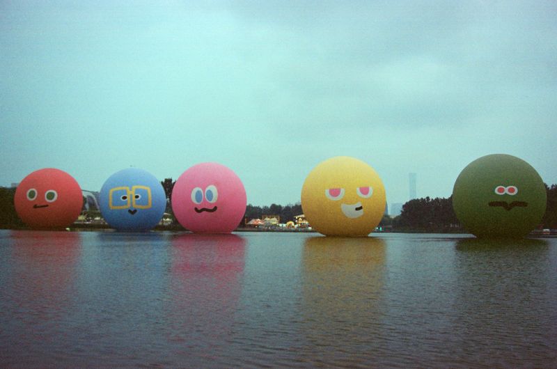Five large, colorful inflatable spheres with cartoon faces floating in a line across a lake under a hazy sky.
