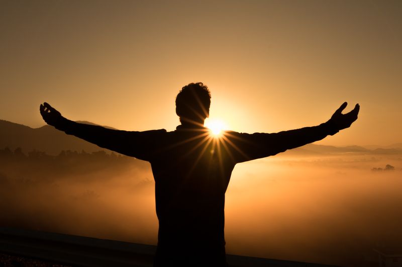A silhouette of a man raising his arms in gratitude while the sun is rising in front of him.