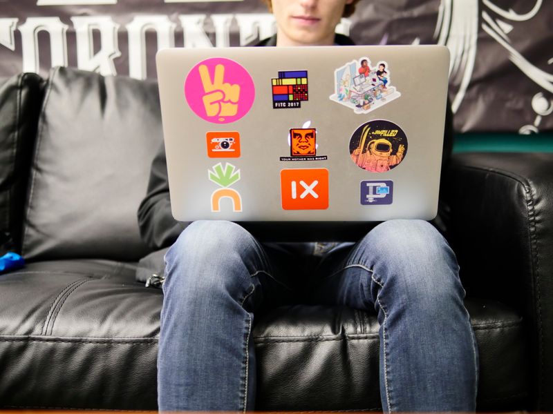 Image: Person in jeans sitting down on a black leather couch with stickers on their laptop.