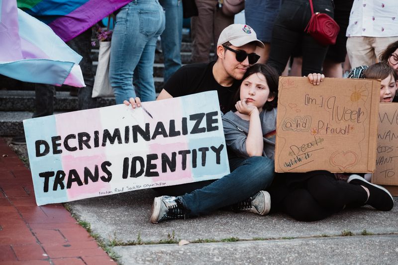 A person sitting on the ground at a protest with a a teenager, holding a sign stating 'Decriminalize Trans Identity'.