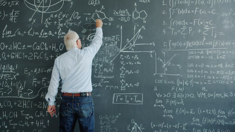 Someone using chalk to write on a blackboard covered in a mix of chemical, math, and physics equations.
