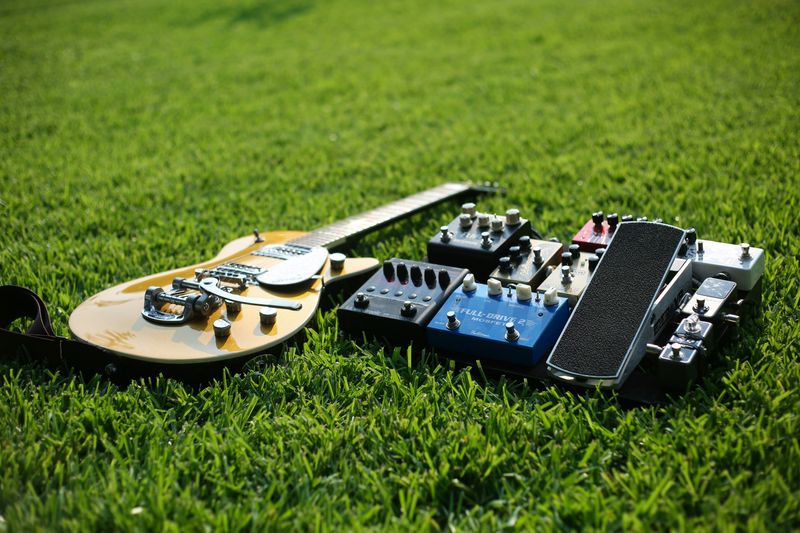 A guitar beside effects pedals in a field of grass.