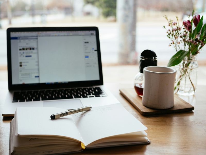 Image of a table with a laptop, open notebook, pen, mug and a vase with some flowers.