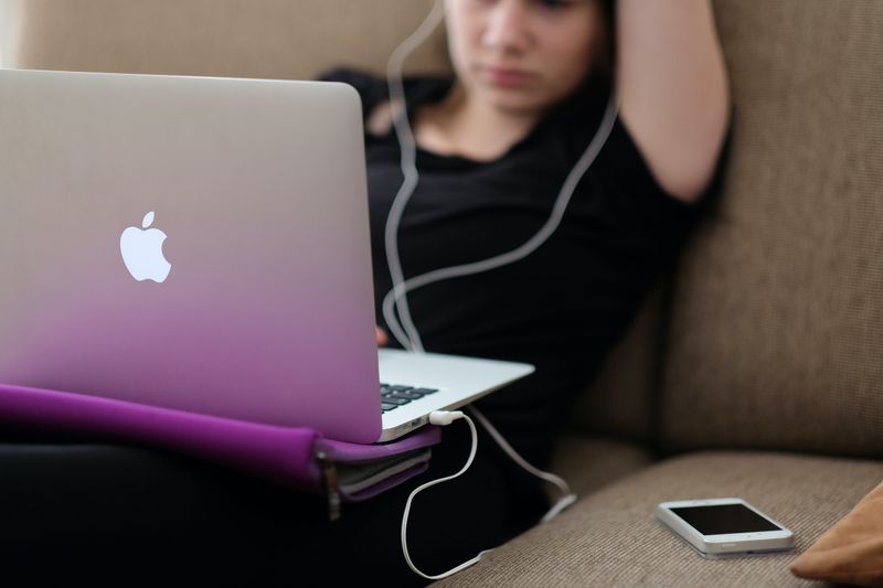 A woman slouching on a couch, working at laptop with headphones and phone nearby.
