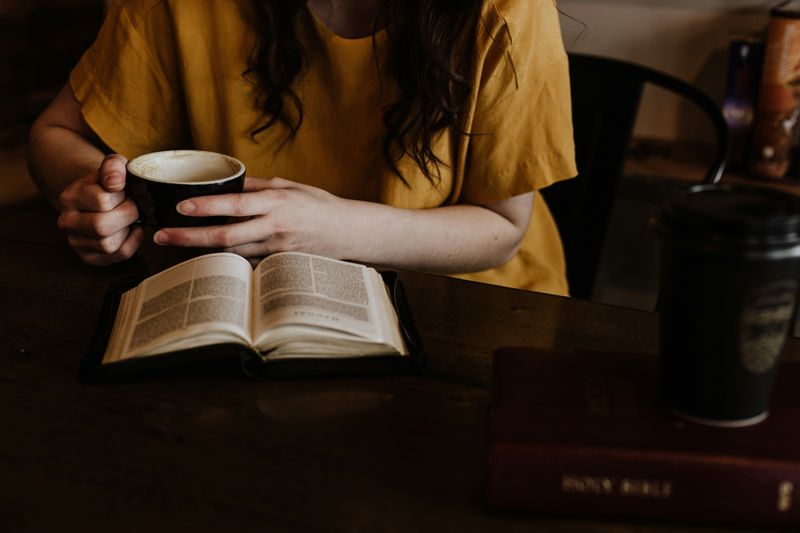 A woman reading a book with a coffee in hand, sitting at a table.