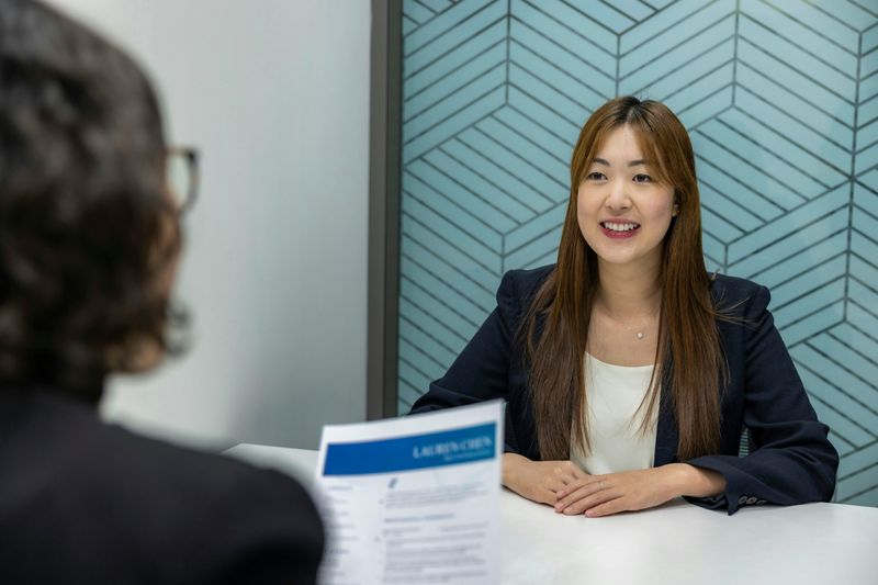 A smiling woman looks across the table at an interviewer, who is holding her resume.