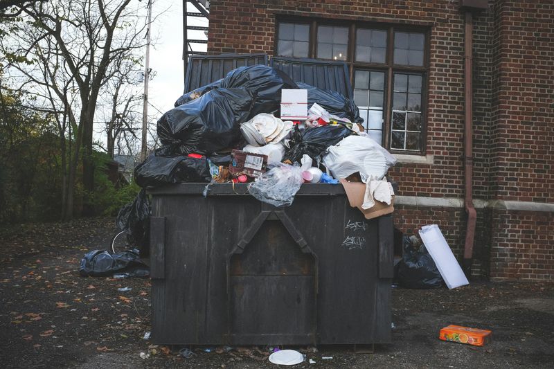 A black dumpster outside of a brick apartment building that is overflowing with black garbage bags and other trash.