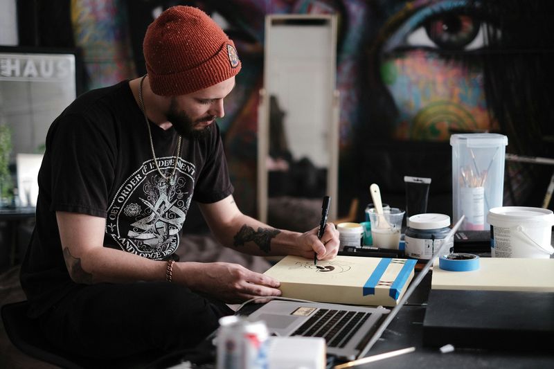 A person sitting at a table drawing on a canvas, with a black marker, beside an open laptop surrounded by artist's materials.