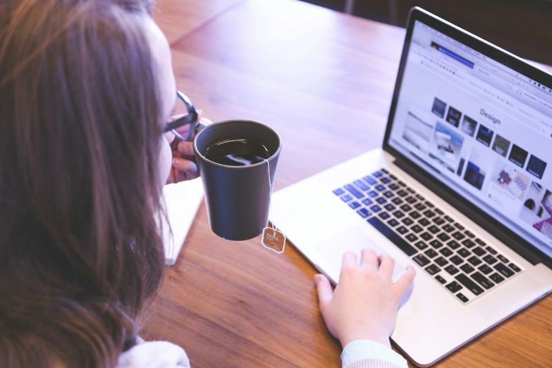 A woman drinking tea while typing on a laptop. She is using AI for website design.