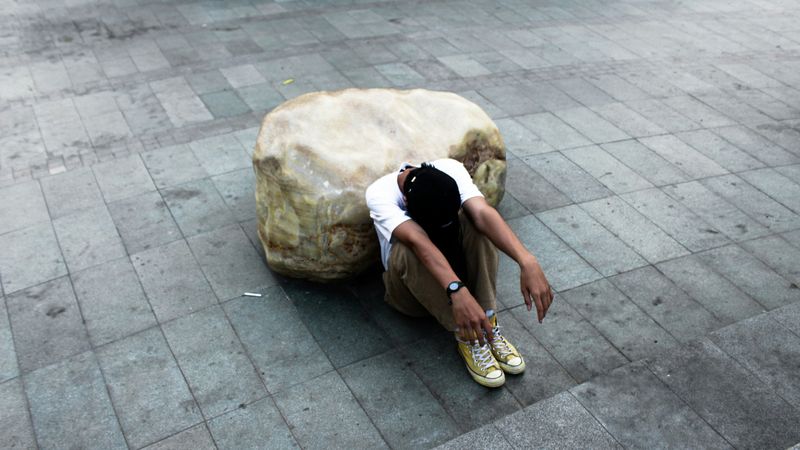 A student is sitting on the ground. His knees are to his chest, his arms are hanging forward, and his head is down.