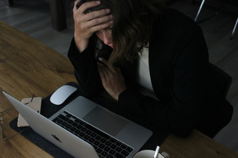 Person hunched over, sitting at a table, holding their head in one hand. They're looking at a laptop in front of them.