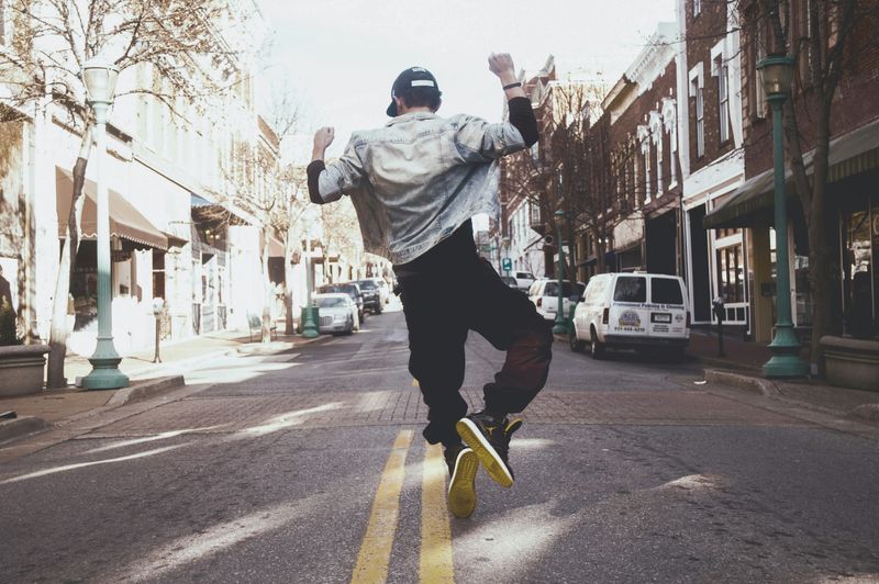 A person jumping up in middle of street, wearing baseball cap, jean jacket, loose pants, and yellow-soled sneakers.