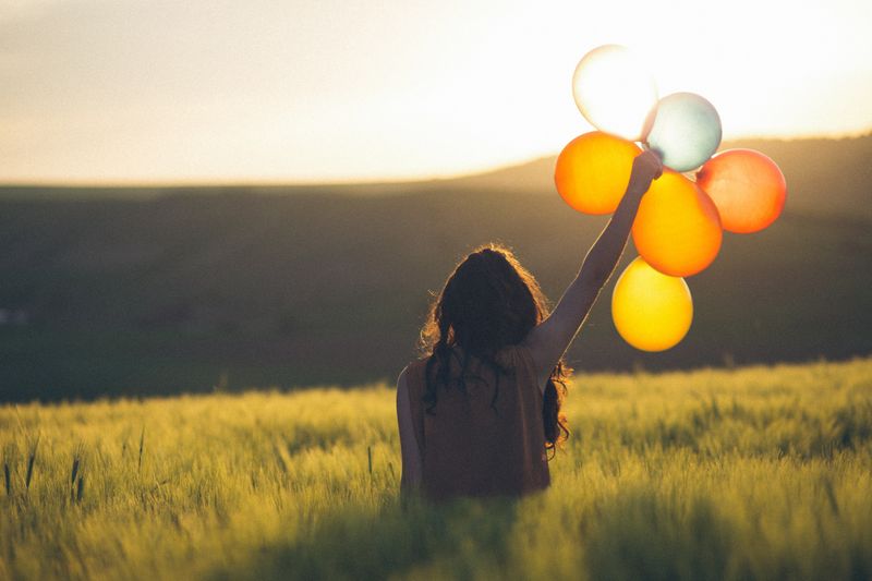 A woman in the grassy fields holds up six balloons toward the sky.