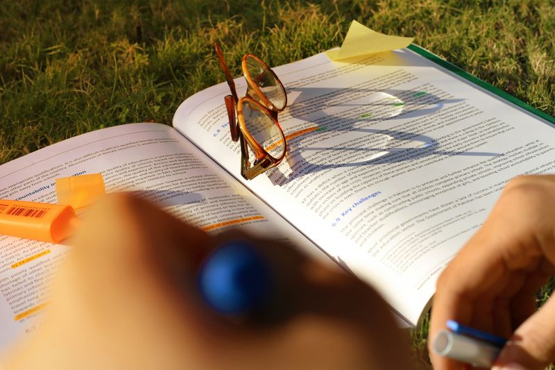 Brown glasses and a highlighter sitting on top an opened textbook on grass.
