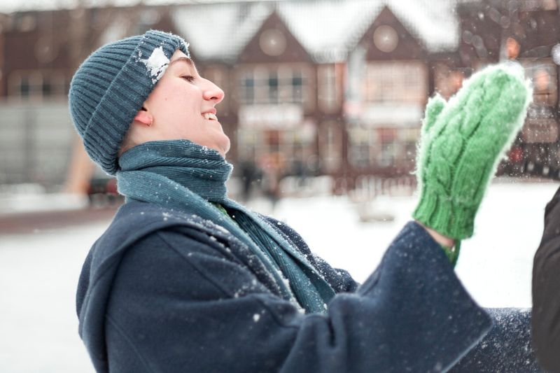 person in the snow wearing a blue jacket, snow hat, scarves, and green gloves with a brown house background 