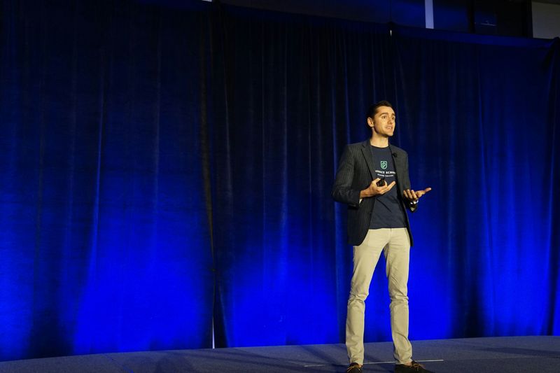 A young man talking on stage with open body language. His palm faces outward and he stands with confident posture.