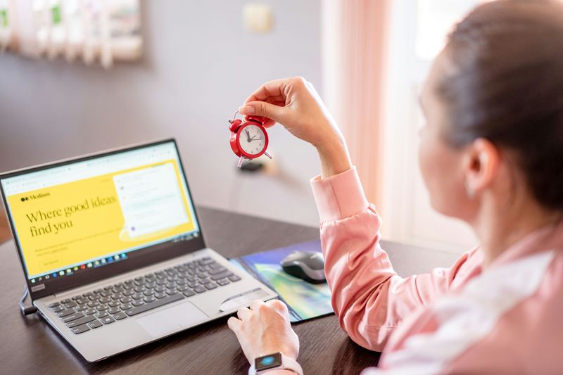 A woman working at a laptop and looking at a small red clock she holds in her right hand.