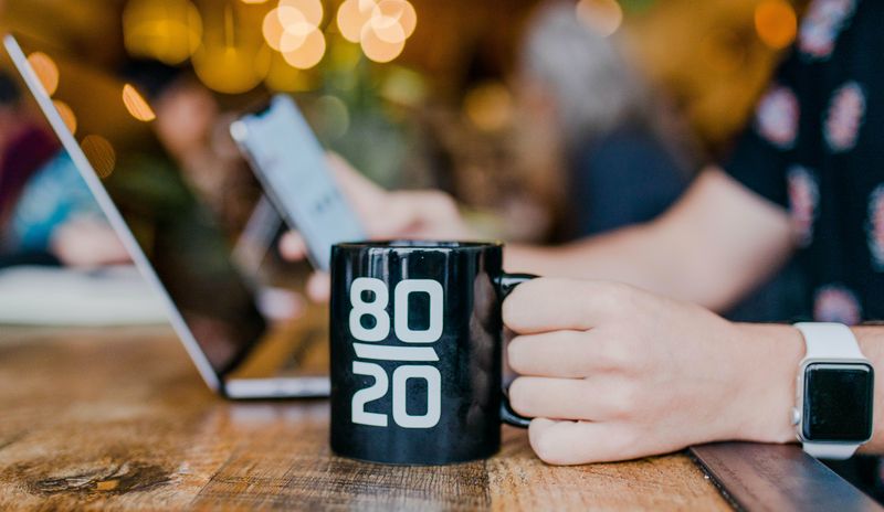 A person holding a mug on a desk. The mug has 80/20 written on it.