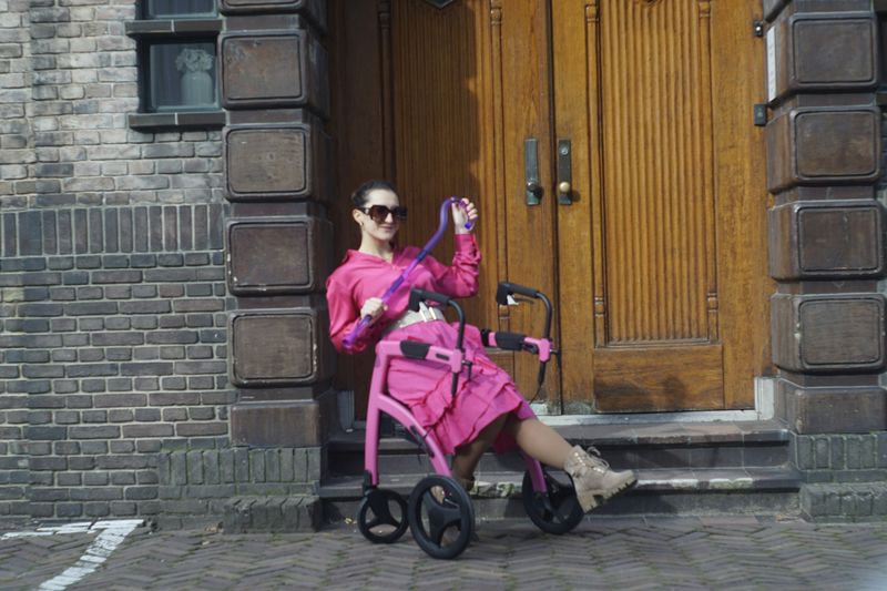 A woman wearing all pink. She sits on a walker outside a door and she holds a flashy cane.