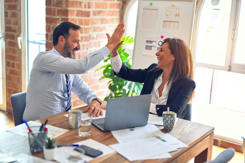 A man and a woman in business attire sitting next to each other, smiling and high-fiving.