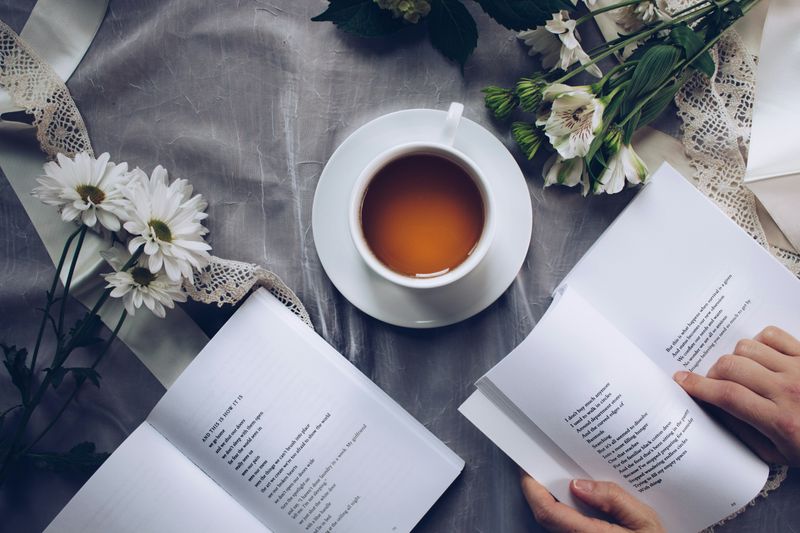 Person reading book of poetry next to flowers and cup of tea