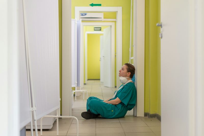 A medical worker in scrubs looking tired and sad as they sit on the floor of a hospital.