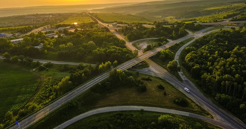 A bird's eye view image of multi-lane highway roads intersecting over each other on a huge green forest during a sunset.