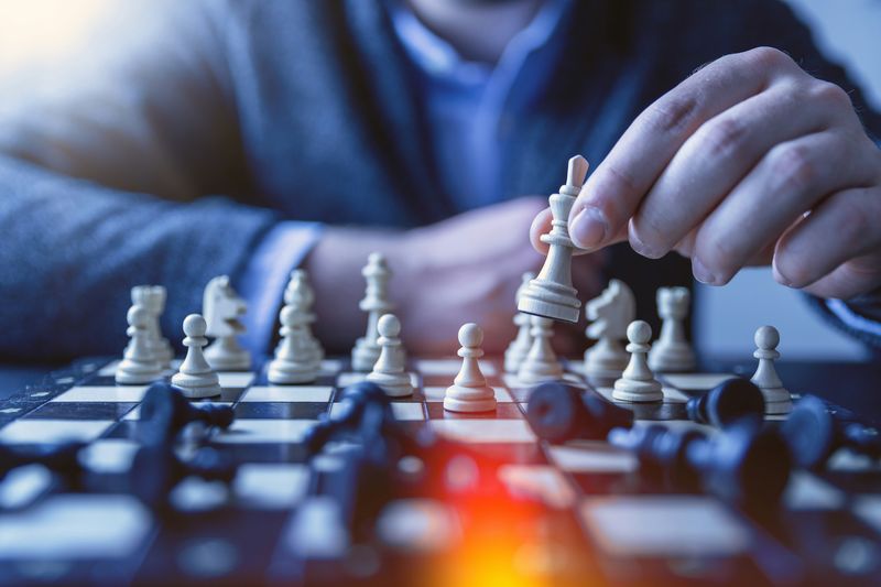 A chessboard and someone's hand holding one of the white pieces. All of the black pieces lie knocked over. 