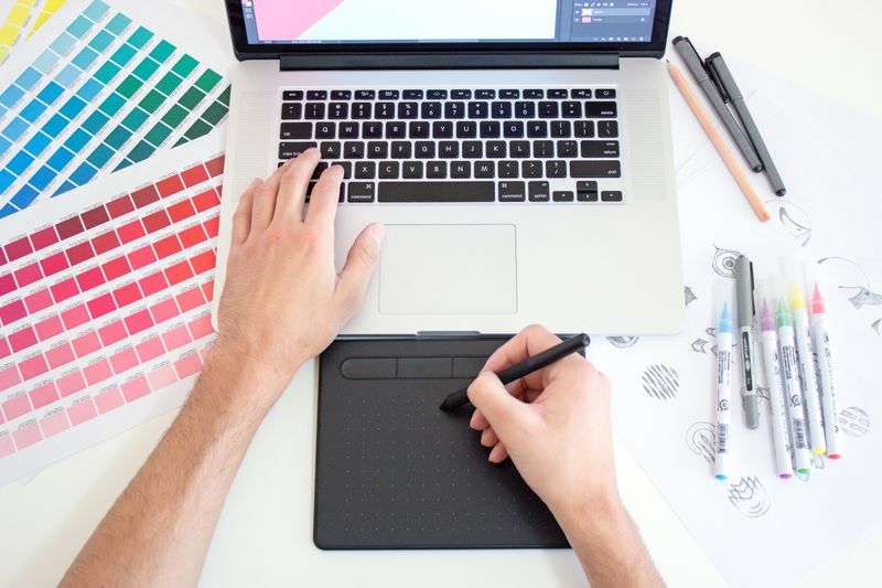 A person designing a poster on a laptop using a drawing pad and color palettes.