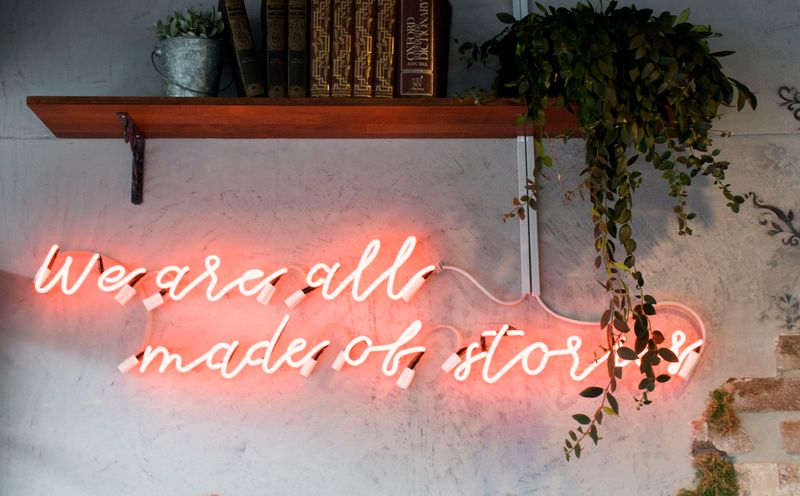 Neon script on the wall under a bookshelf - the script says 'we are all made of stories'