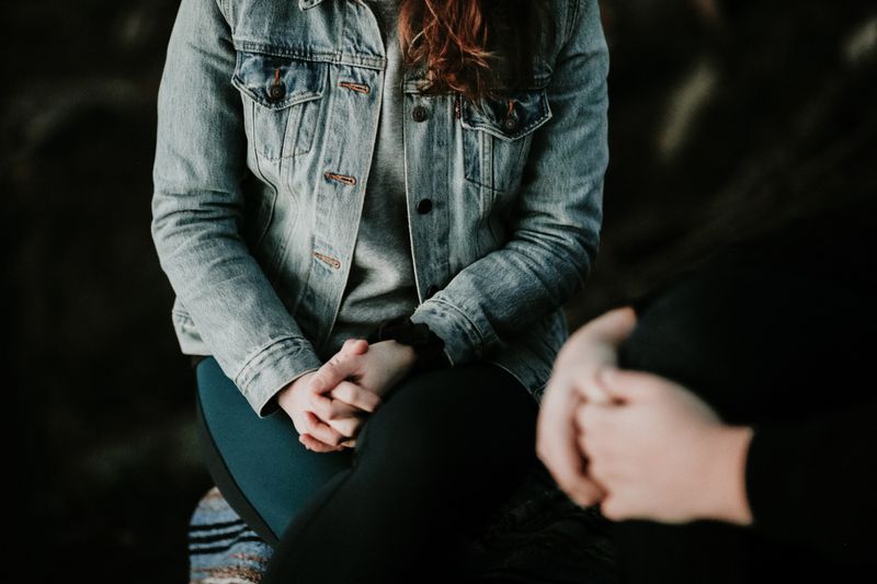 Person wearing a denim jacket, hands clasped, facing towards another person whose hands are also clasped.