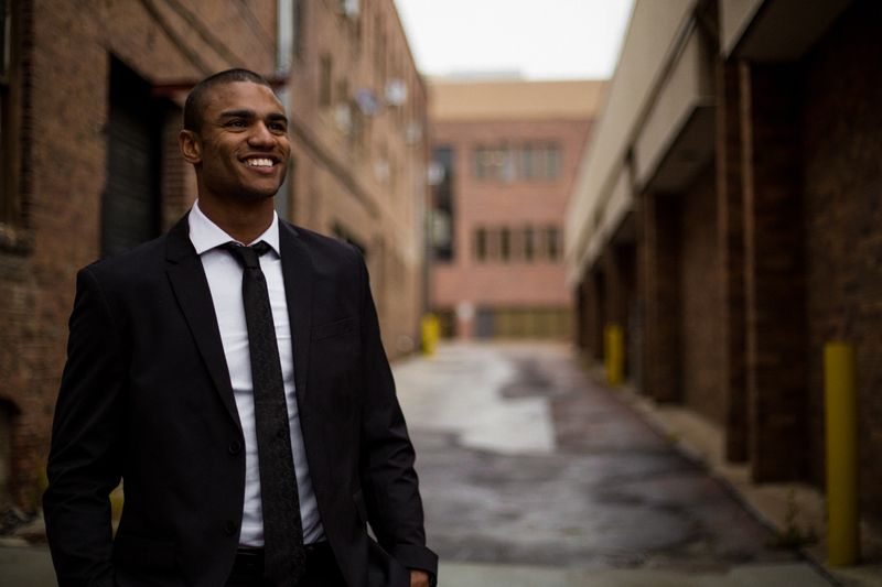 Young smiling man wearing a suit with a blurred brick building background.