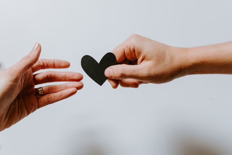 A hand giving a black paper heart to another outreached hand.