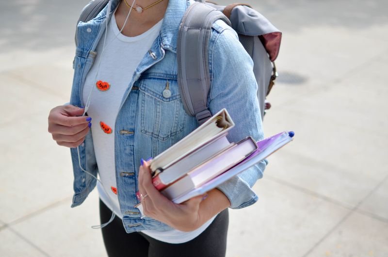 A student wearing a white t-shirt and denim jacket is carrying a stack of books and wearing a backpack.