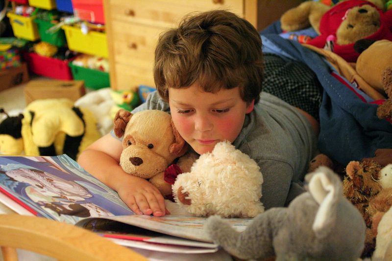 Young boy laying in bed with stuffed animals reading a picture book